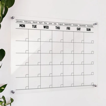Clear acrylic modern calendar with bold MON-SUN headers and blank monthly grid, mounted beside a plant