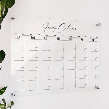 Clear acrylic 'Family Calendar' with script title and rounded monthly grid, mounted on a wall beside a plant