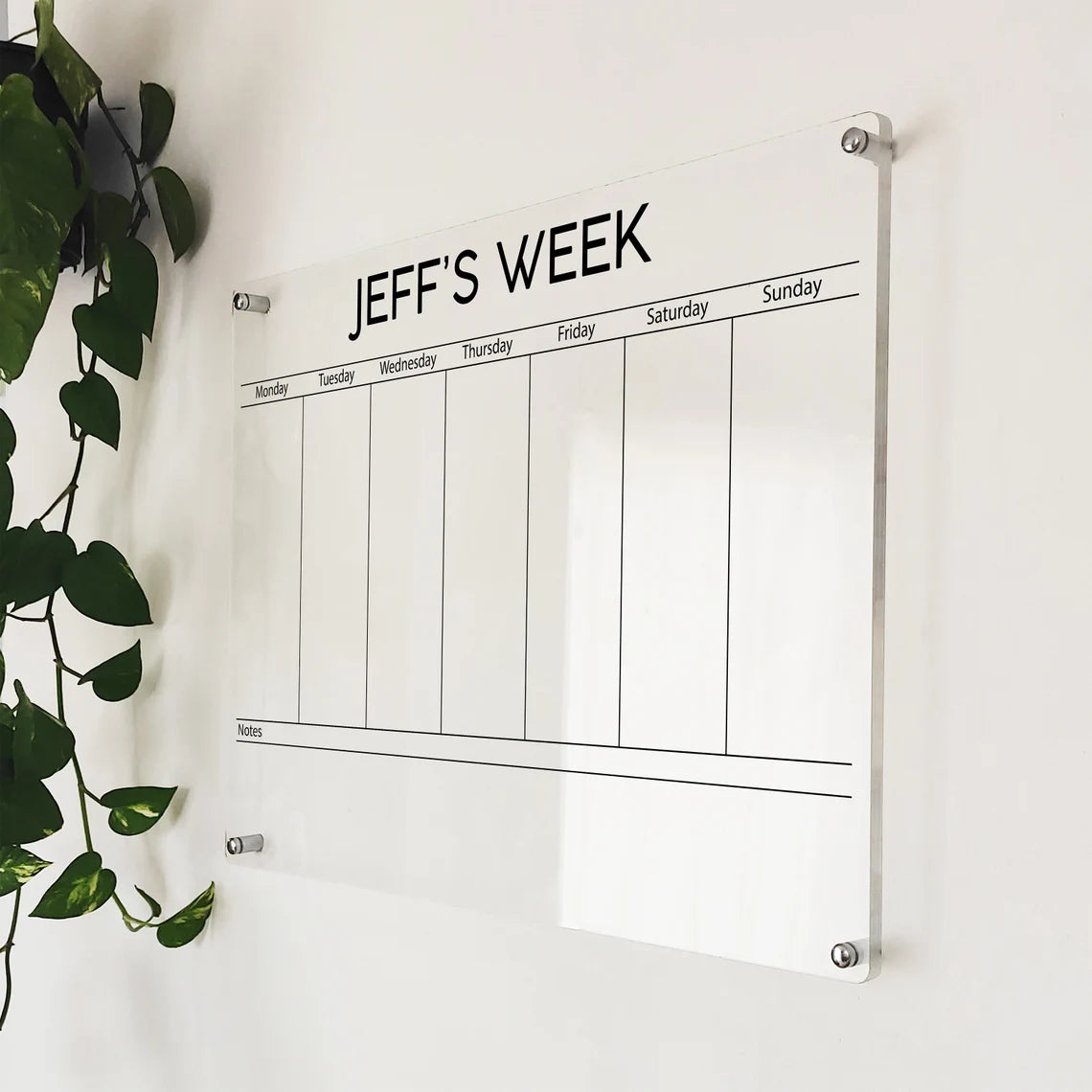 Angled close-up of the personalized 'Jeff's Week' acrylic weekly planner mounted beside a hanging plant