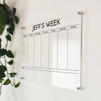 Angled close-up of the personalized 'Jeff's Week' acrylic weekly planner mounted beside a hanging plant