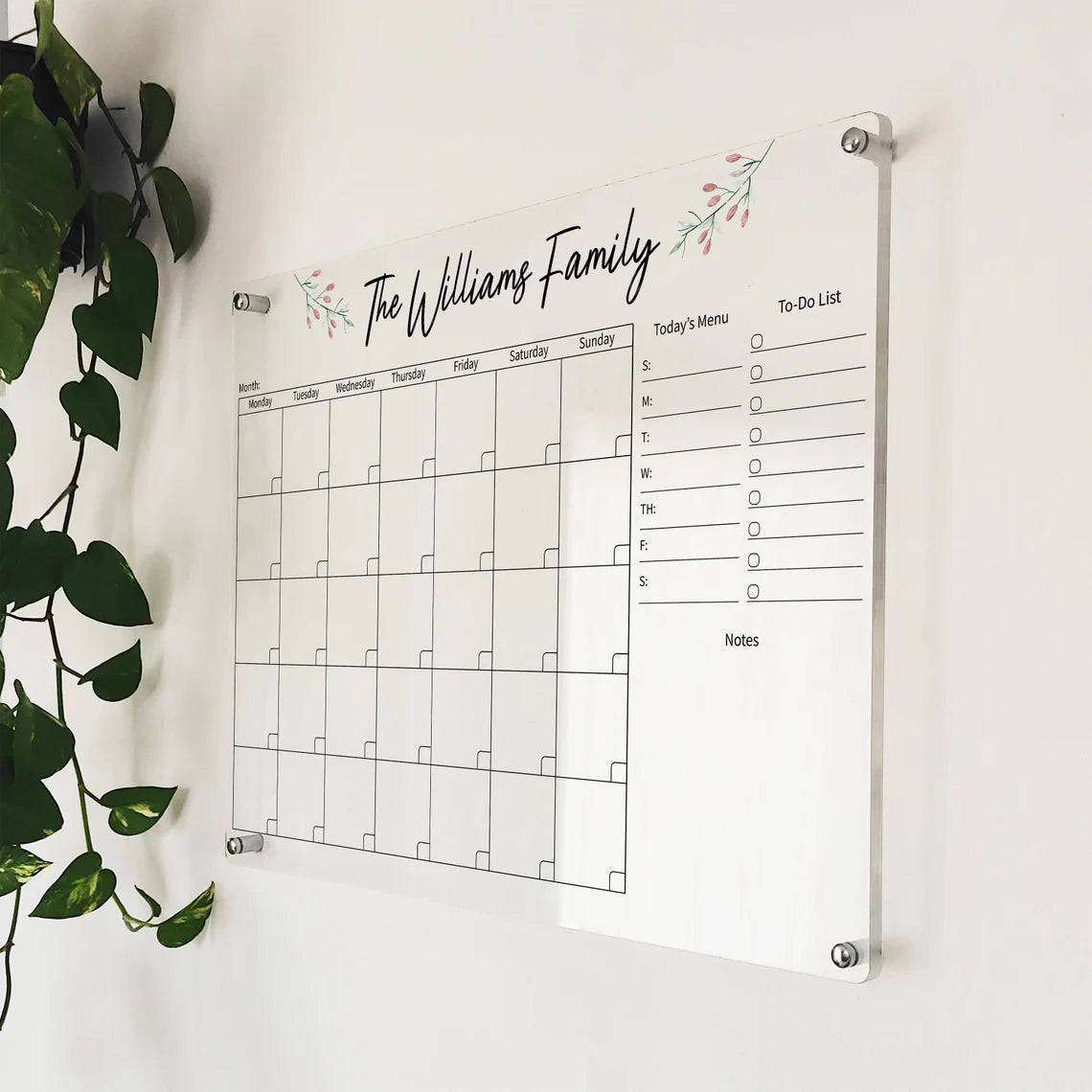 Angled close-up of 'The Williams Family' floral acrylic calendar mounted beside a hanging plant