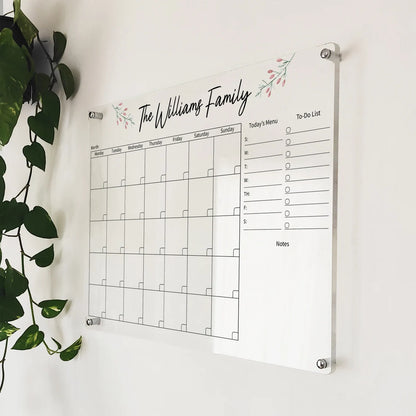 Angled close-up of 'The Williams Family' floral acrylic calendar mounted beside a hanging plant