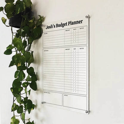 Angled close-up of 'Josh's Budget Planner' acrylic income and expense tracker beside a hanging plant