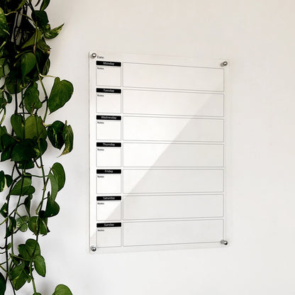 Clear acrylic weekly planner with black day-of-week headers and note boxes, mounted beside a plant