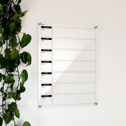 Clear acrylic weekly planner with black day-of-week headers and note boxes, mounted beside a plant