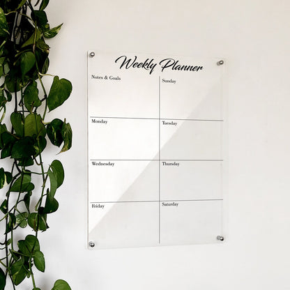 Clear acrylic 'Weekly Planner' with script title, notes and goals block, and 4x2 day grid, mounted beside a plant