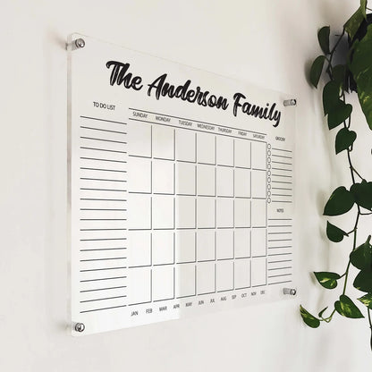 Angled close-up of 'The Anderson Family' acrylic calendar with grocery and to-do lists beside a plant