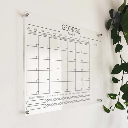Angled close-up of the 'George Family' acrylic calendar with habit tracker mounted beside a plant