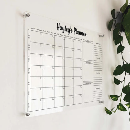 Angled close-up of 'Hayley's Planner' acrylic calendar with appointments and goals mounted beside a hanging plant