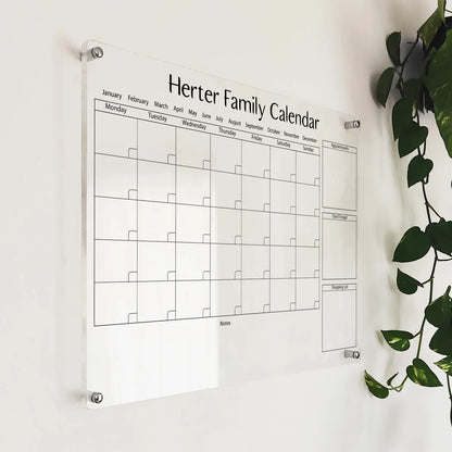 Angled close-up of the 'Herter Family Calendar' acrylic planner mounted beside a hanging plant
