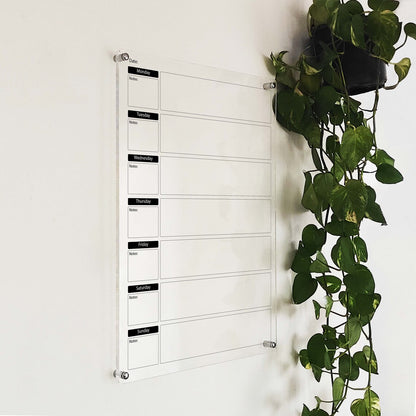 Angled close-up of the acrylic weekly dry-erase planner with day tabs, mounted near a hanging plant