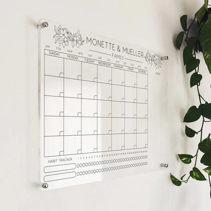 Angled close-up of the floral-accented family acrylic calendar with habit tracker beside a plant