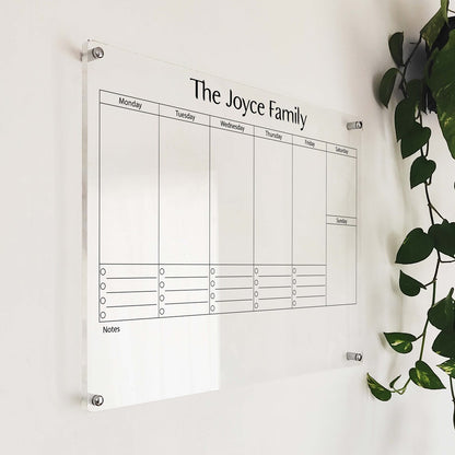 Angled close-up of 'The Joyce Family' acrylic weekly planner mounted beside a hanging plant