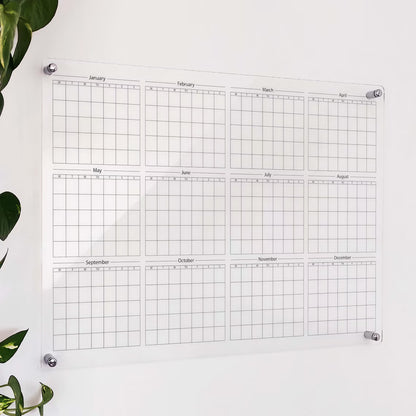 Clear acrylic yearly wall calendar with 12 blank monthly grids, mounted on a wall beside a plant