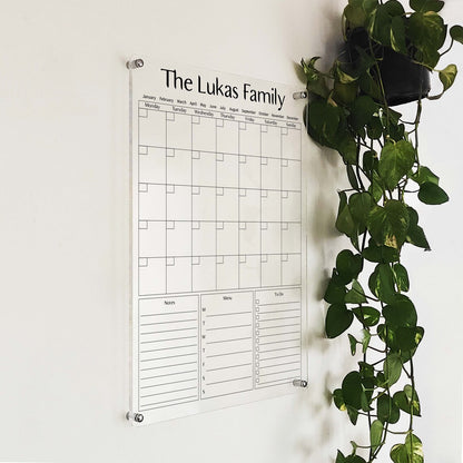 Angled close-up of the 'The Lukas Family' acrylic calendar mounted beside a hanging plant