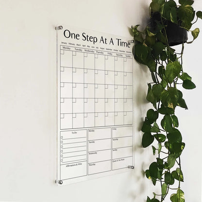 Angled close-up of the 'One Step At A Time' acrylic calendar and affirmations board beside a hanging plant