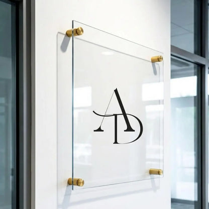 Angled close-up of a clear acrylic 'AD' monogram sign held by brass standoff bolts